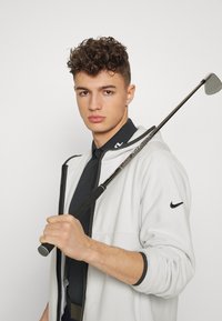 Osoba trzyma kij golfowy, ubrana w czarną koszulę z kołnierzykiem i białą kurtkę zapinaną na zamek z szarym wnętrzem i czarnymi akcentami.
