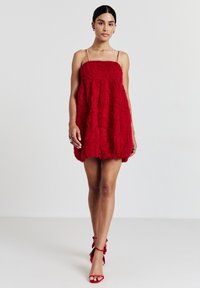 Frau im roten strukturierten Mini-Kleid mit Spaghettiträgern und passenden roten hochhackigen Sandalen, vor einem schlichten weißen Hintergrund stehend.