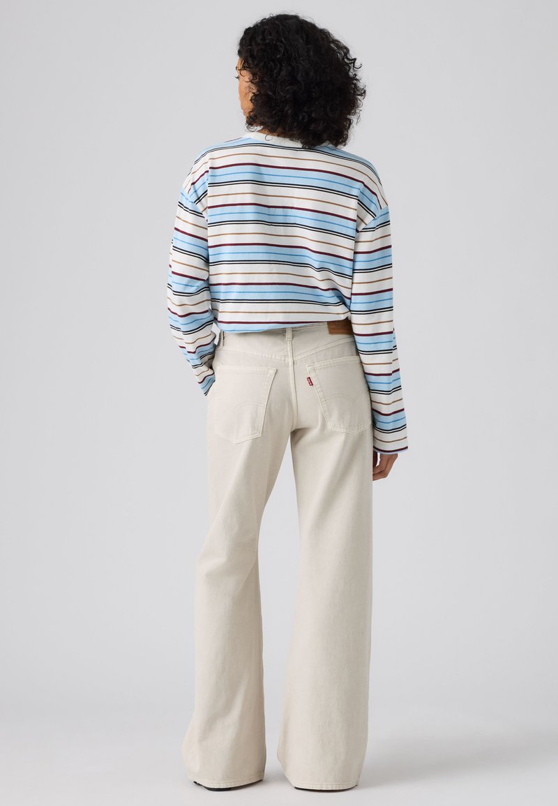 Personne aux cheveux bouclés portant un haut rayé à manches longues et un pantalon crème taille haute à jambes larges, debout de dos devant un fond uni.