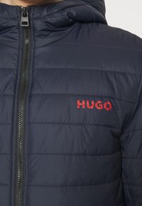 Marinblå pufferjacka med horisontell sömnad, blixtlås och röd "HUGO"-logotyp på vänster sida. Har även huva och en slät yta.
