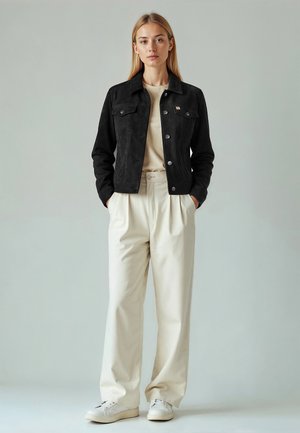 Ung kvinde står med hænderne i lommerne iført sort knappet jakke, beige skjorte, cremelange bukser med vide ben og hvide sneakers mod grå baggrund.