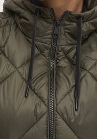 Zbliżenie na oliwkowozieloną pikowaną kurtkę z czarnym zamkiem błyskawicznym i sznurkami do regulacji przy kołnierzu.