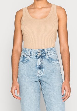 Persoon in een beige geribbelde mouwloze top gestoken in een hooggeknipte lichtblauwe denim jeans met dubbele knoopsluiting.