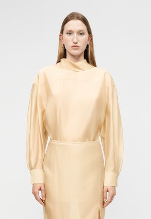 Γυναίκα με ίσια ξανθά μαλλιά φοράει φόρεμα μπεζ σατέν με μακριά μανίκια και ψηλό γιακά, στέκεται μπροστά σε ένα απλό λευκό φόντο.