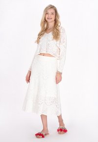 Hvid hæklet crop top med en flared hvid nederdel, der har et paisley mønster. Modellen bærer lyserøde og sølvbelagte slidesandaler.