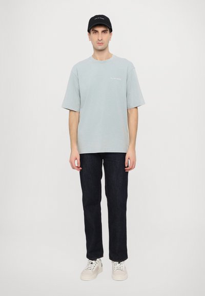 T-shirt oversize in cotone azzurro chiaro con maniche corte, abbinata a jeans in denim scuro e sneakers bianche con lacci, completata da un cappellino nero.