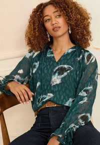 Blusa teal con stampa astratta bianca e nera, maniche lunghe con dettagli a volant e scollatura a V. Abbinata a jeans scuri e una cintura marrone.