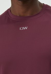 Nærbilde av en person som har på seg en bordeaux sportsskjorte med rund hals og reflekterende "ICIW"-logo midt på brystet.