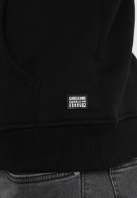 Zwarte hoodie met ribbelrand en manchetten, voorzien van een klein label met de tekst "CARSJEANS SUPPLY GOODS 82" erop genaaid aan de zijkant.