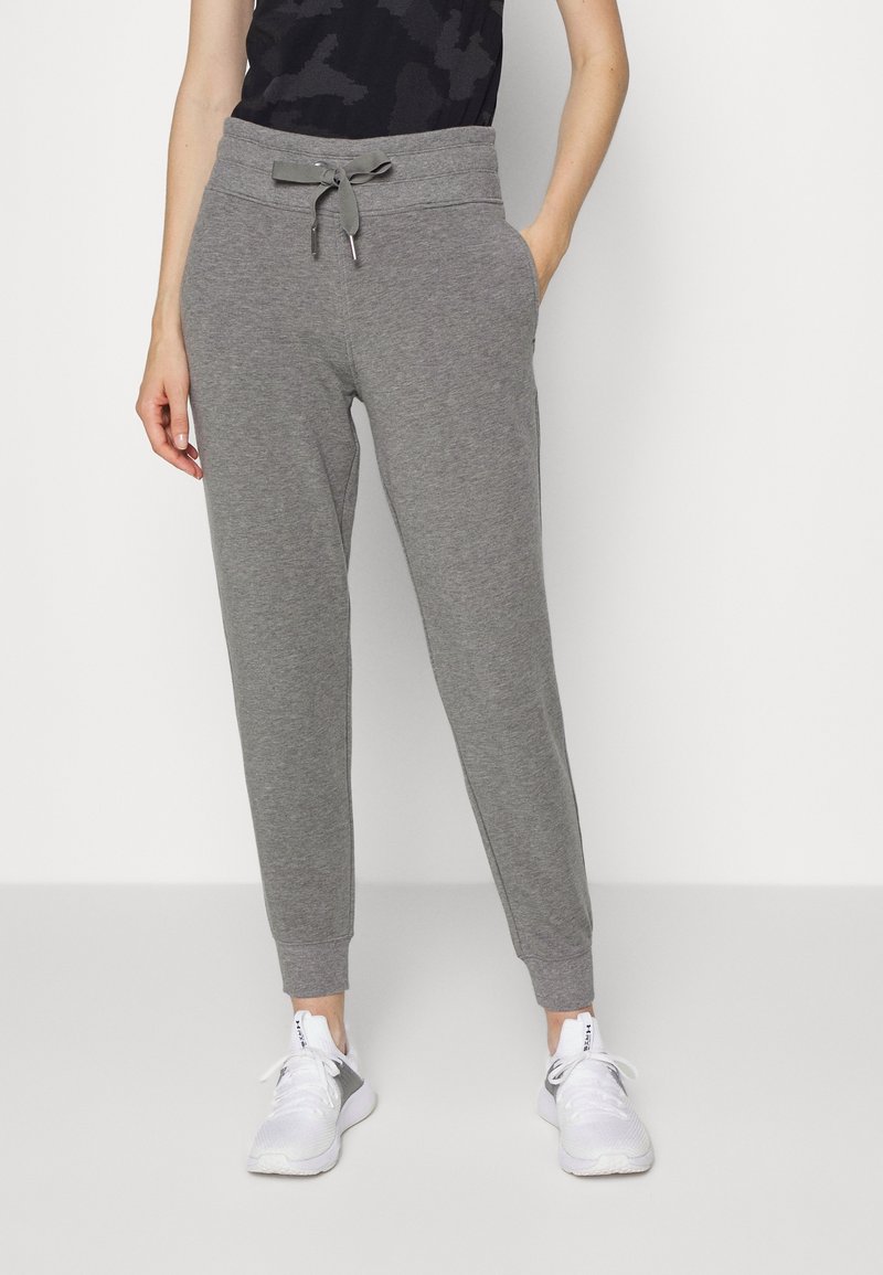 Femme portant un pantalon de jogging gris avec cordon de serrage et des baskets blanches, debout les mains dans les poches sur un fond blanc uni.