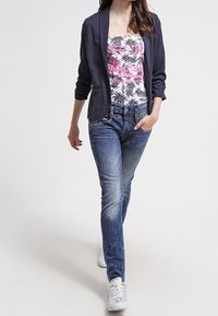 Žena v námořnickém blejzru, bílé grafické tílko s růžovým a černým vzorem, modrých džínách a bílých teniskách, kráčející s jednou rukou v kapse.