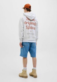 Le hoodie gris présente un grand imprimé "Édition Studio Originals" au dos, associé à un short en denim bleu et des bottes beiges. La casquette est orange.
