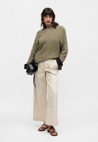 Γυναίκα που φοράει πουλόβερ πλεκτό σε ελιά χρώμα, μαύρο πουκάμισο με γιακά, φαρδιά μπεζ παντελόνια, καφέ σανδάλια, κρατώντας μαύρη τσάντα, στέκεται μπροστά σε λευκό φόντο.