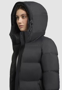 Schwarze Pufferjacke mit großer Kapuze, ausgestattet mit einem gesteppte Design, Reißverschluss und minimalem Branding auf der Vorderseite. Glatte Textur.