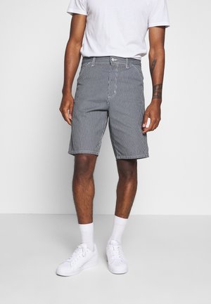 Mand iført sort-hvide lodrette stribede shorts, hvid t-shirt, hvide sokker og hvide sneakers, stående mod en ensfarvet baggrund.