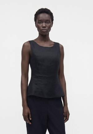 Femme aux cheveux courts et bouclés portant un haut noir sans manches près du corps et un pantalon noir, debout devant un fond clair uni.