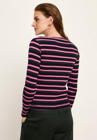 Frau mit nassen, nach hinten gegelten Haaren, die goldene Creolen trägt und ein langärmeliges Oberteil mit geripptem, pinkem und schwarzem horizontalem Streifenmuster sowie schwarze Hosen anhat.