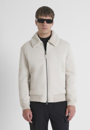 Veste bomber beige avec un col en fausse fourrure, fermeture éclair et poignets côtelés. Portée sur une chemise noire, accompagnée de lunettes de soleil noires.