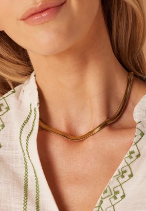 Gouden ketting met een glad, gebogen ontwerp. Draagt om de nek en contrasteert met een linnen kledingstuk met groene borduursels.
