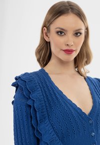 Maglione blu lavorato a maglia con scollo a V profondo, spalle arricciate e dettagli con bottoni. La texture presenta motivi in rilievo su tutta la superficie.