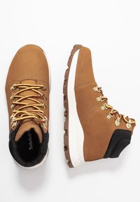 Bruna lädervandringsskor med svarta accenter, gula och röda skosnören, gyllene öglor och en strukturerad vit mellansula. Robust gummisula.