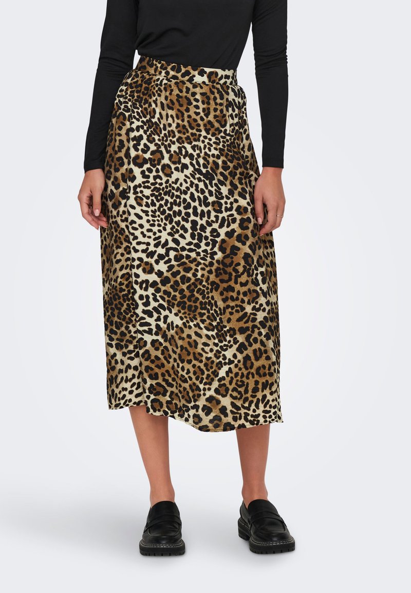 Donna che indossa una gonna midi con stampa leopardata, un top nero a maniche lunghe e scarpe slip-on nere, in piedi davanti a uno sfondo semplice.