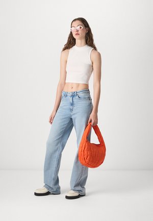 Sac en tissu matelassé orange tenu dans une main, associé à un haut sans manches à côtes blanc et un jean large bleu clair, avec des chaussures contrastantes.