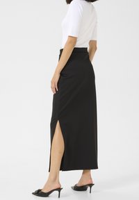 Jupe maxi stretch noire avec une haute fente, design ajusté, texture lisse, associée à des sandales à talons noires et un haut blanc.