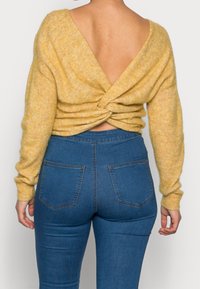 Geel gebreid truitje met een diepe V-hals aan de achterkant en gedraaide details bij de taille. Gepaard met high-waisted blauwe spijkerbroek met achterzakken.