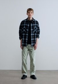 Μαύρο και άσπρο καρό φανελένιο πουκάμισο πάνω από μαύρο t-shirt, συνδυασμένο με ανοιχτό πράσινο παντελόνι cargo και μαύρα αθλητικά παπούτσια, σε ουδέτερο φόντο.