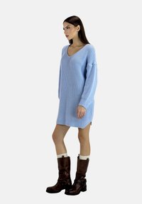 Abito maglione azzurro chiaro con scollo a V e maniche lunghe. Presenta un motivo a texture e spacchi laterali, abbinato a stivali marroni al ginocchio.