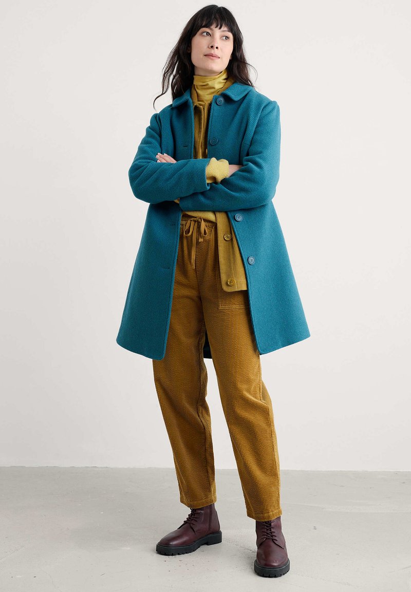 Femme aux cheveux foncés portant un manteau bleu-vert, un col roulé moutarde, un pantalon en velours côtelé moutarde et des bottes en cuir lacées marron foncé, se tenant les bras croisés.