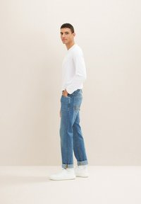 Bela srajca z dolgimi rokavi in modri jeans z zavihanimi hlačnicami, v kombinaciji z belimi supergami. Preprost dizajn, brez vzorcev ali vidnih logotipov.