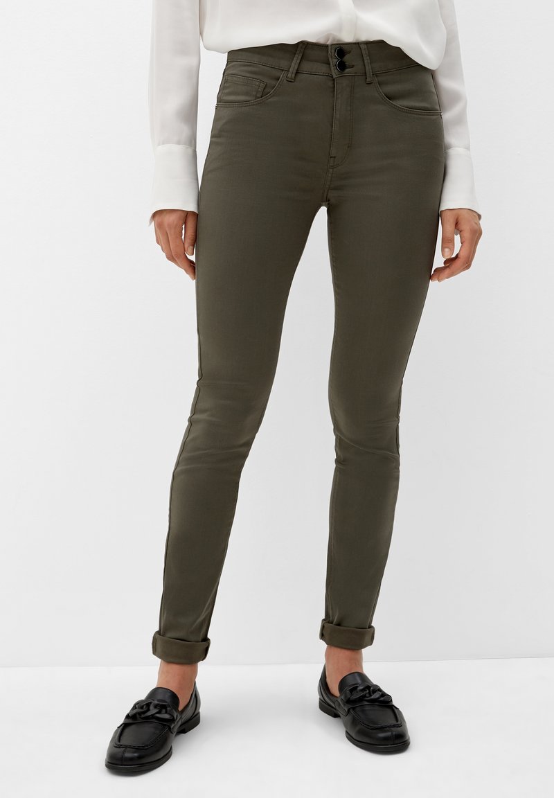 Personne portant un pantalon skinny kaki foncé retroussé, une chemise blanche à manches longues et des mocassins noirs, debout devant un fond uni.
