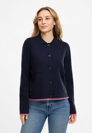 Frau trägt eine marineblaue Strickjacke mit Knöpfen, rosa Besatz und zwei Brusttaschen, kombiniert mit blauen Jeans, stehend vor einem einfachen Hintergrund.