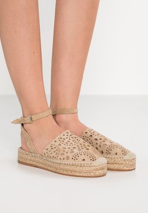Fødder iført beige espadrillesandaler med perforerede overdele og ankelsnore, stående på et hvidt gulv.