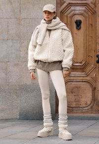 Kremowa kurtka bomber z grubym splotem na swetrze, noszona na beżowym golfie, w połączeniu z lekkimi legginsami i kremowymi futrzanymi butami z prążkowanymi mankietami.