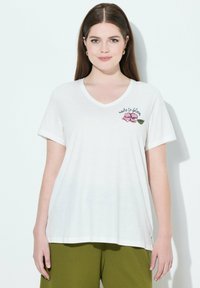 Camiseta blanca de cuello en V, hecha de tela ligera, con bordado de una flor rosa y una hoja verde, acompañado del texto "hecha para florecer". Mangas cortas.