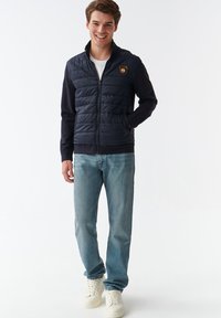 Mørkeblå puffer-vest med lynlås foran, quiltet tekstur og bjørne-logo på brystet; parret med lyseblå jeans og hvide sneakers.