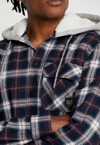 Navy plaid shirt with gray hood, featuring black buttons, two chest pockets, and a soft interior lining; intricate check pattern with red accents.
