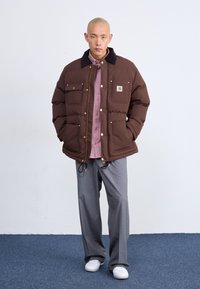 Chaqueta tipo puff en color marrón con detalles en dorado y dos bolsillos frontales, llevada sobre una camiseta de color rosa claro. Los pantalones grises y las zapatillas blancas completan el conjunto.
