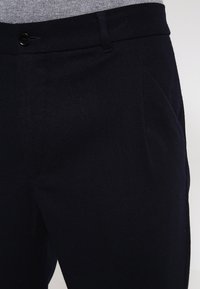 Tmavě modré kalhoty z texturované látky s knoflíkovým zapínáním, předními záhyby a klasickým střihem. Bez viditelných kapes.