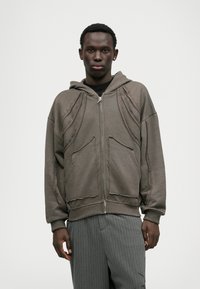 Grå lynlåsjakke med slidte kanter, store forlommer og hætte. Har tekstureret sømdetaljer langs skuldrene.