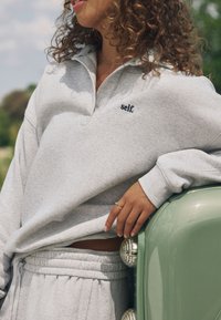 Hall hallide kaelusega dressipluus, millel on tumesiniselt tikitud "self." logo. Tekstuuriga kangas, kombineeritud sobivate hallide dressipükstega.