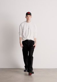 Λευκό φούτερ με μικρό κόκκινο λογότυπο, μαύρο παντελόνι και μαύρο καπέλο με κόκκινη λεπτομέρεια. Μοντέλο που περπατά σε ουδέτερο φόντο.