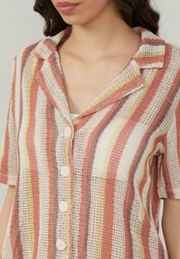 Multicolore gestreepte shirt gemaakt van open geweven stof; heeft een kraag en zes knopen aan de voorkant. Kleuren zijn koraal, geel en room.