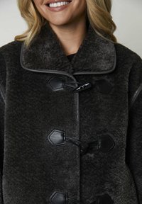 Gros plan d'une personne souriante portant un manteau en peluche gris foncé avec des attaches en cuir noires et un large col.