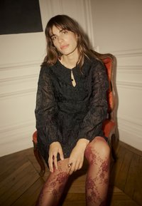 Rochie neagră din dantelă cu mâneci lungi și umflate și detaliu în formă de cheia la gât, purtată cu ciorapi din dantelă burgundy cu modele florale.