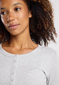 Graues, geripptes Langarmshirt mit Rundhalsausschnitt und sechs Knöpfen. Natürliche Haare, die in sanften Locken gestylt sind, umrahmen den Kragen.