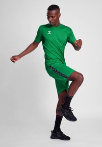Grøn træningsskjorte og shorts med sorte detaljer, der har et logo på brystet. Kompositmaterialer, glat tekstur, båret med sorte sneakers.